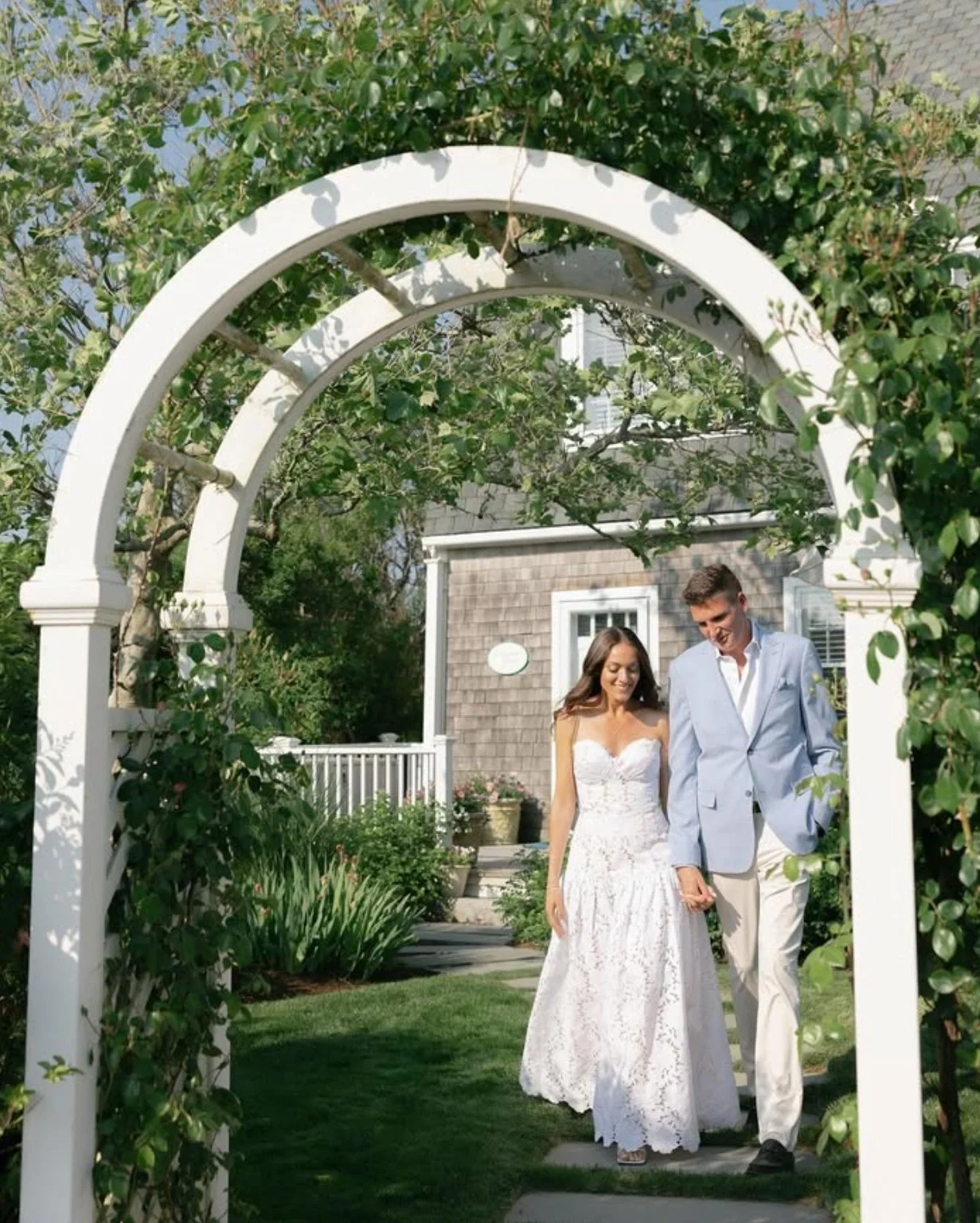 A couple walking under a white garden arch with greenery, a woman in a white wedding dress and a man in a light blue suit, smiling, outdoors in a garden setting.