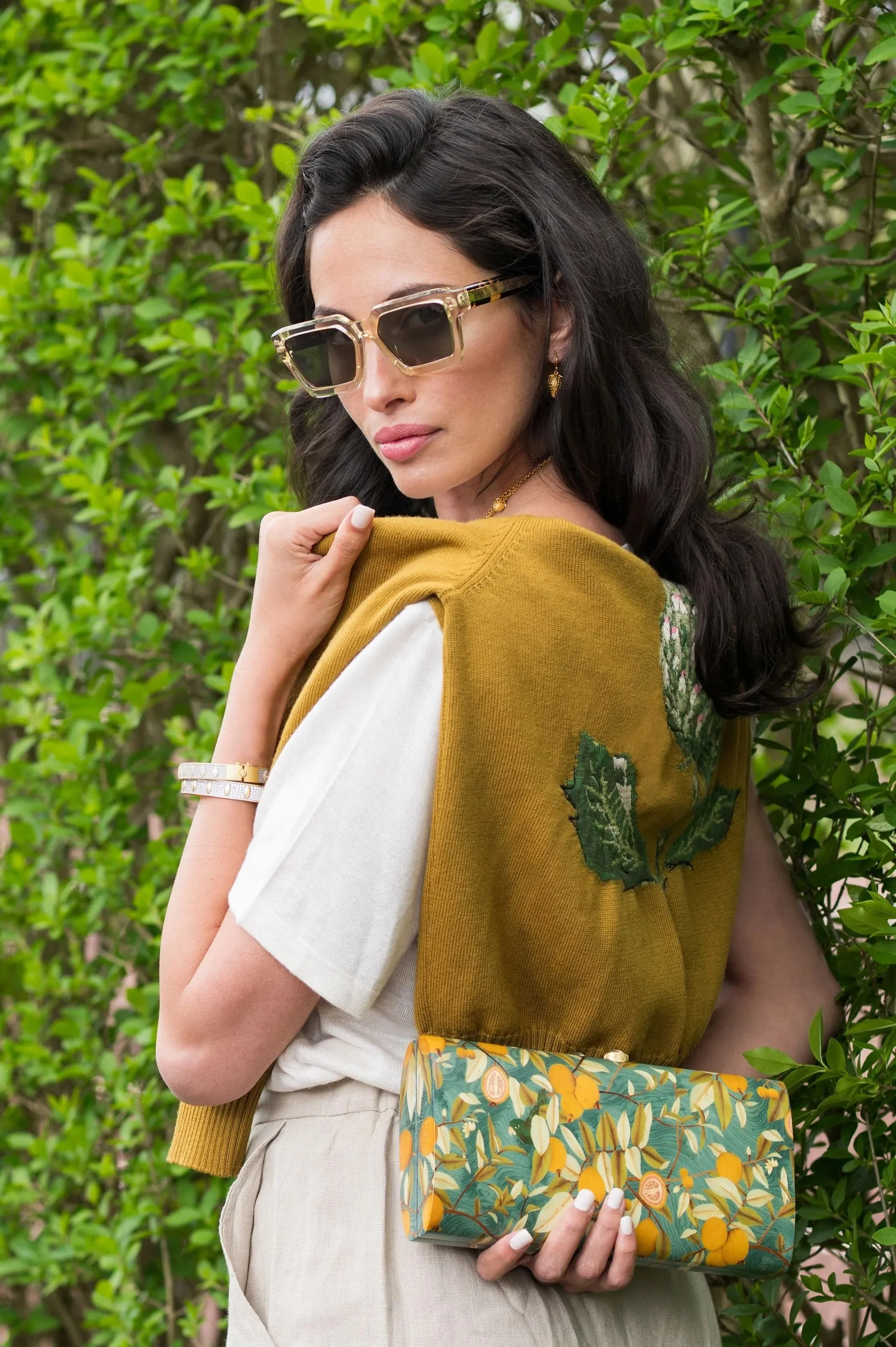 A woman with dark wavy hair and wearing large sunglasses, a white shirt, beige pants, a mustard yellow vest with floral embroidery, holding a colorful clutch with a lemon and leaf pattern, standing in front of a green leafy background.