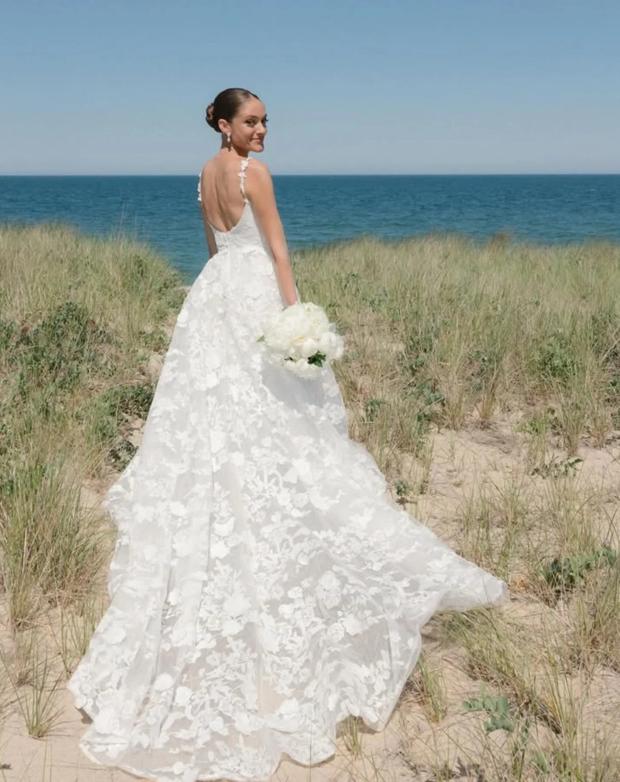 A bride in a white wedding dress holding a bouquet standing on a sandy beach with green grass, with the ocean and clear blue sky behind her.