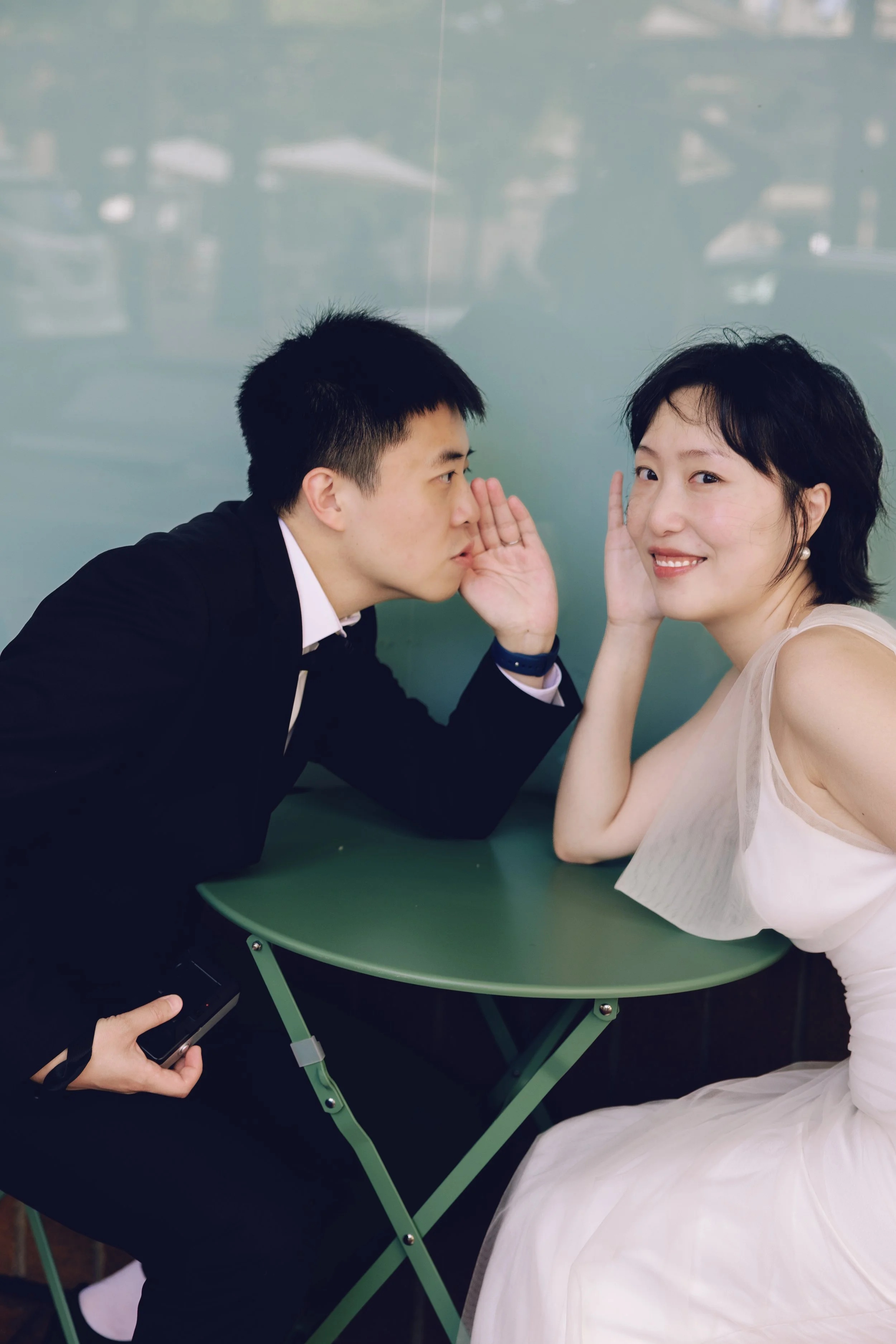 Playful engagement portrait of a couple seated at a small green table.
