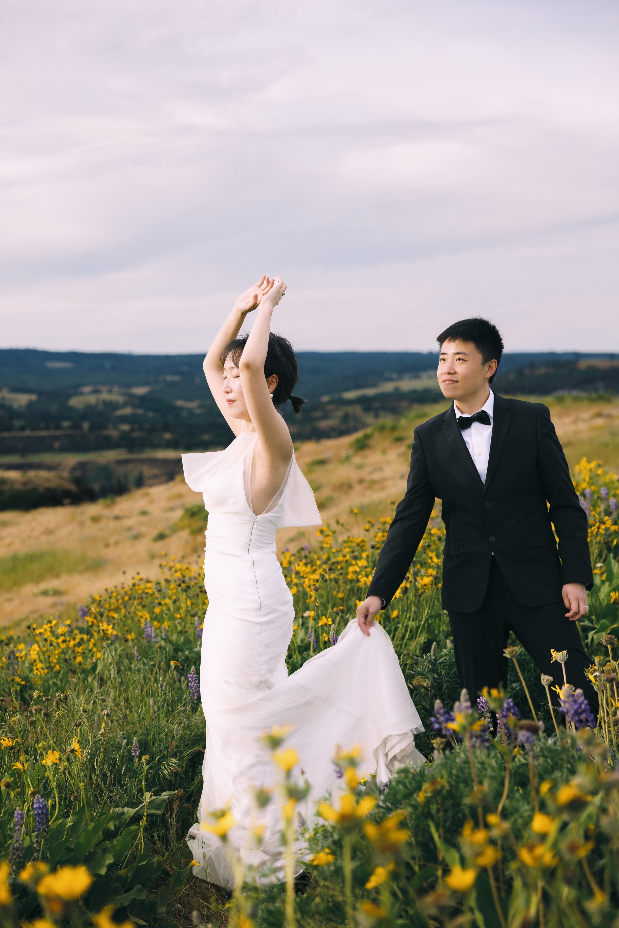 Couple standing among spring wildflowers, with the groom holding the bride’s dress as she lifts her arms.