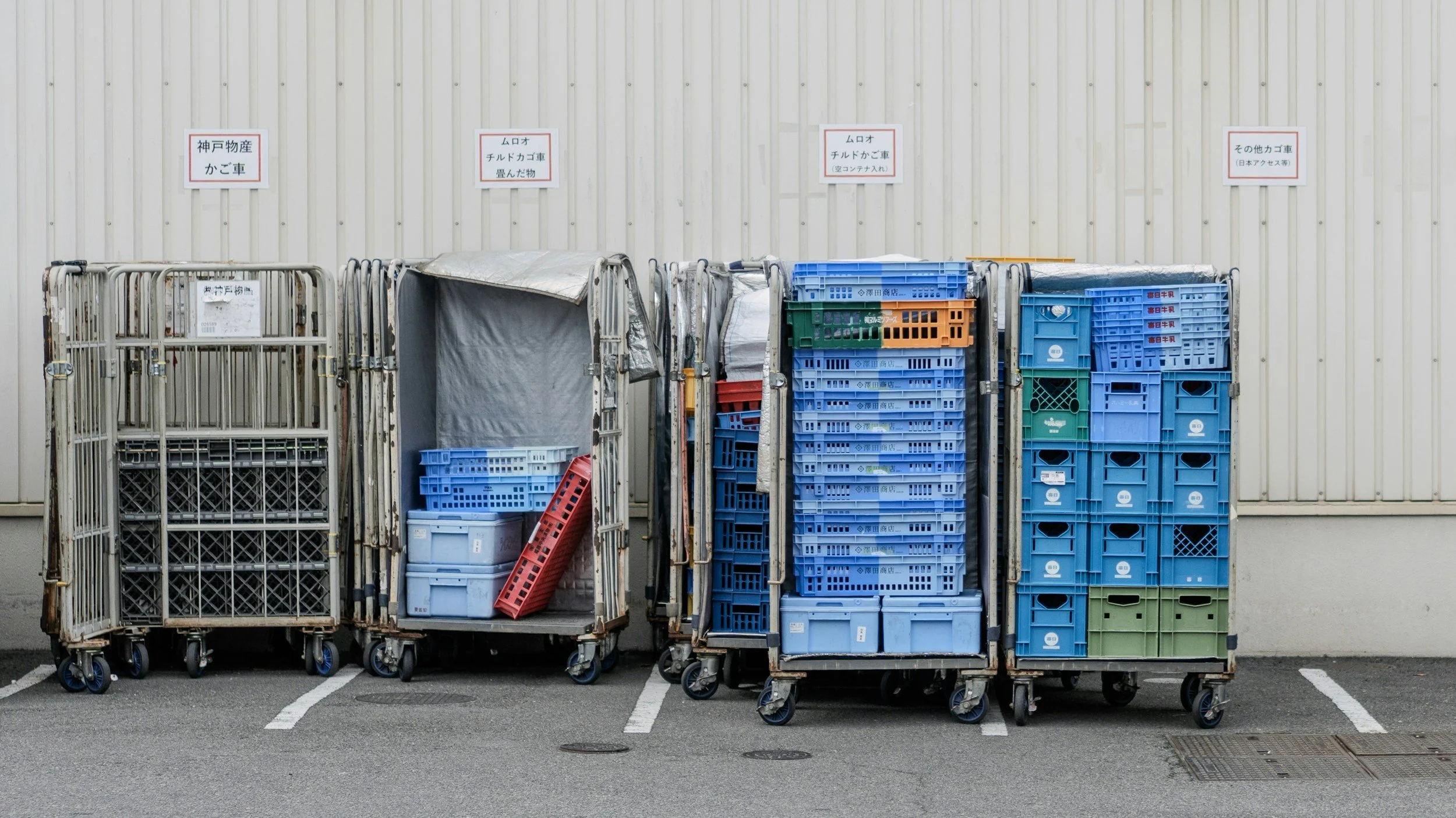 Four carts filled with plastic crates and containers are parked in a lot against a beige corrugated metal wall with signs in Japanese above each cart.