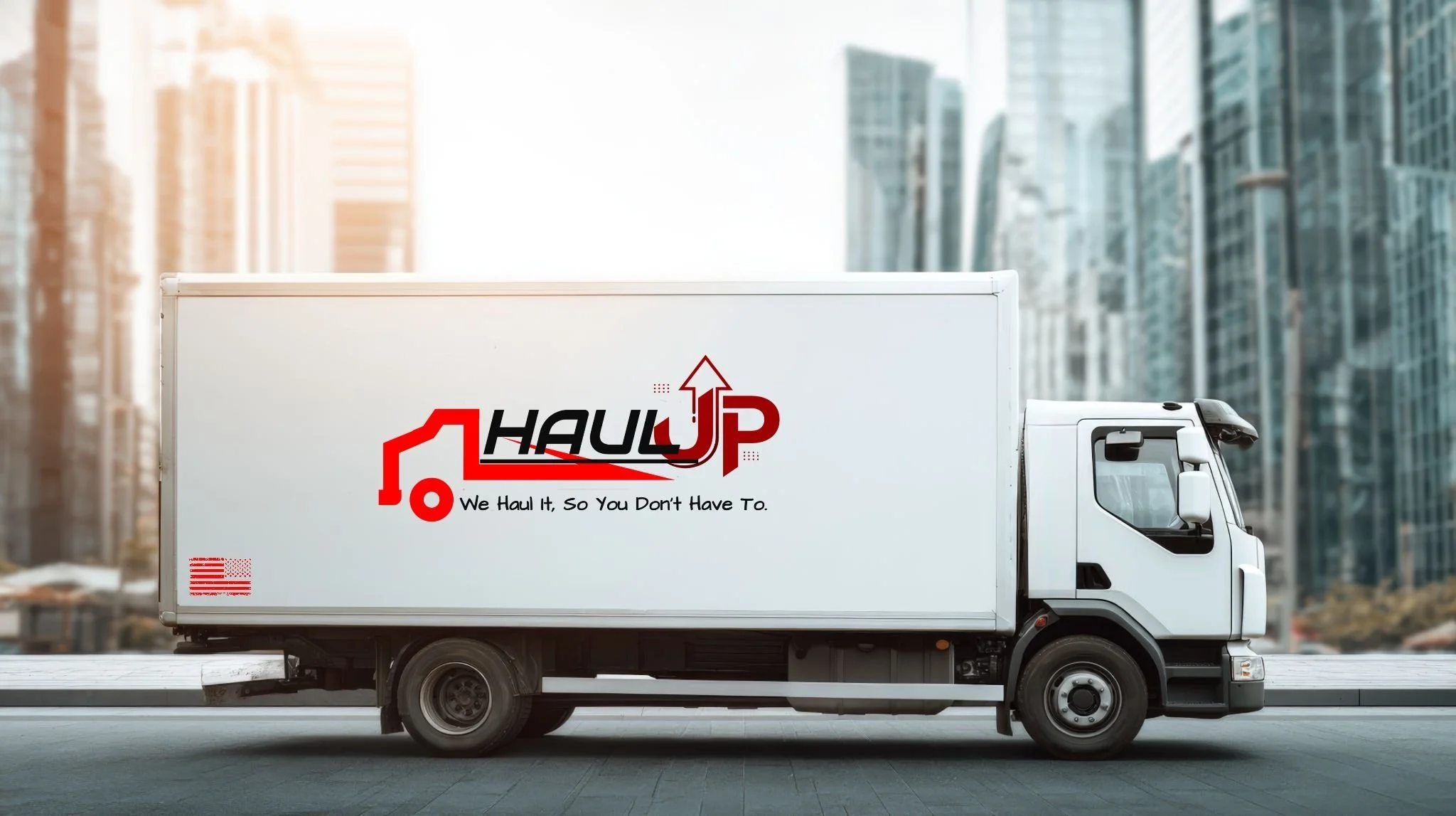 A white moving truck with a logo on the side that says "HAUL UP" and a slogan "We Haul It, So You Don't Have To." The background shows a cityscape with tall glass buildings.