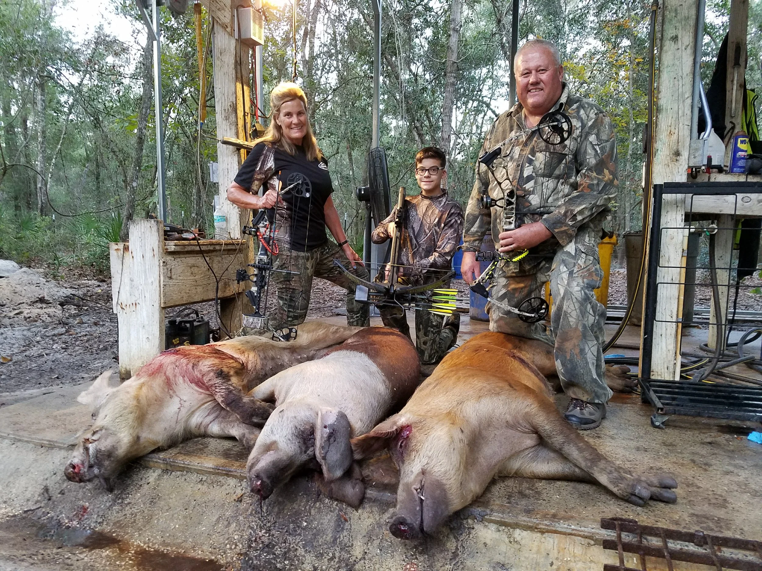 Three people posing with two dead boars on the ground, holding bows, in an outdoor wooded area.