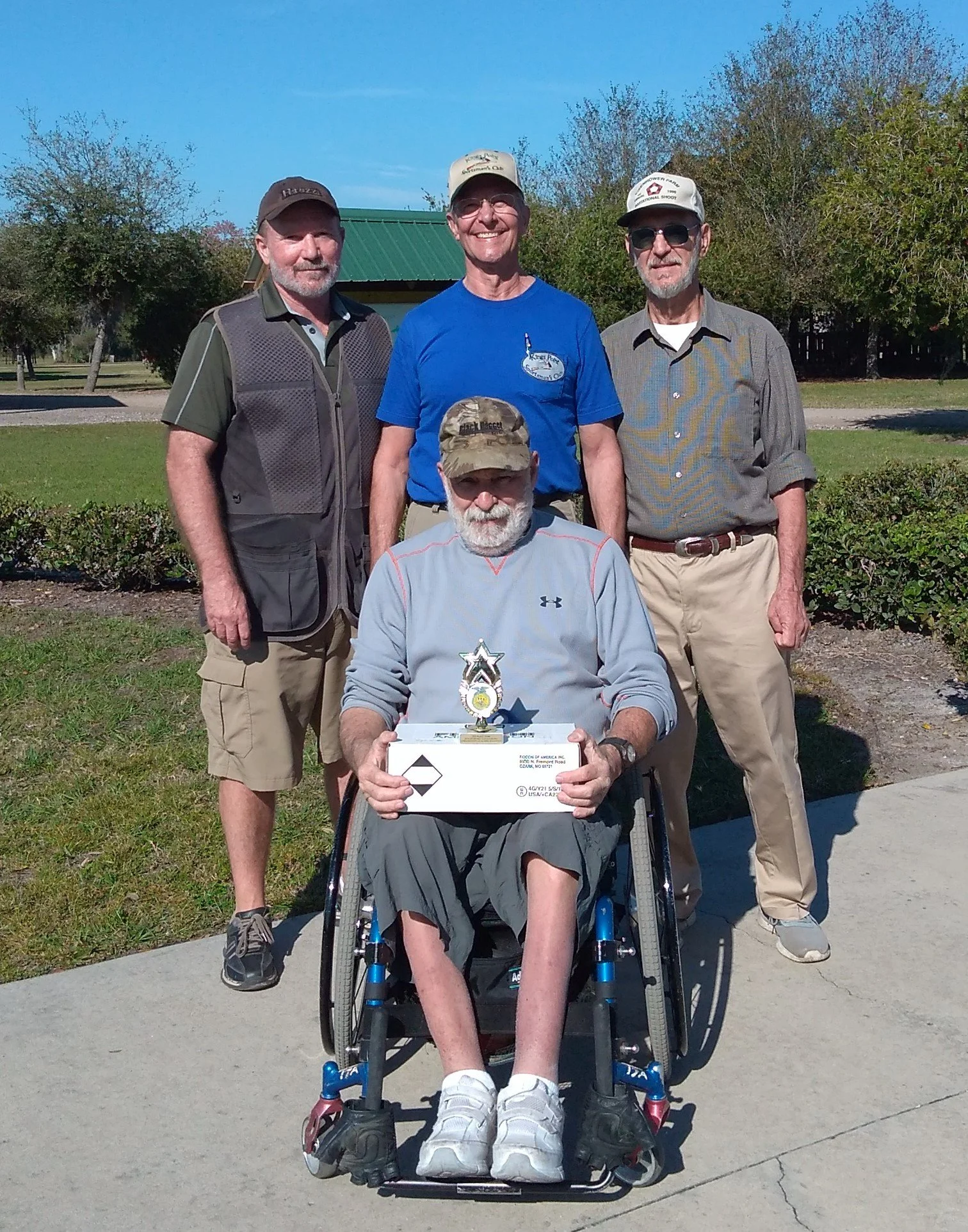 Five men posing outdoors on a sunny day, one in a wheelchair holding a trophy and a box, surrounded by four standing men, with trees and a blue sky in the background.