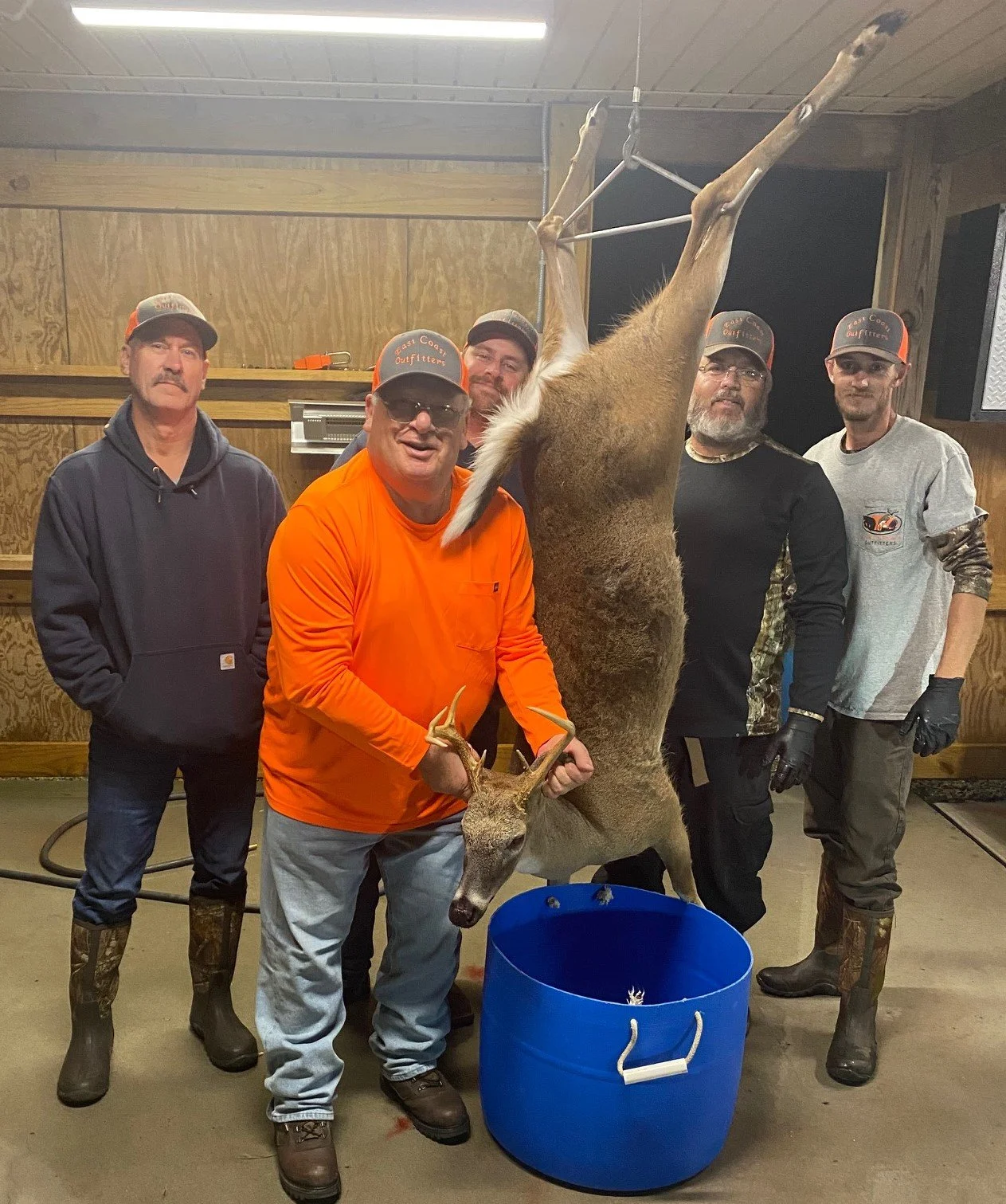 Five men standing inside a wooden shed, posing with a freshly hunted deer hanging upside down. The men are wearing outdoor gear. One man in orange is holding the deer's antlers.
