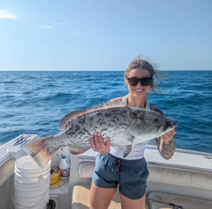 Woman on a boat holding a large fish she caught, with the ocean in the background.