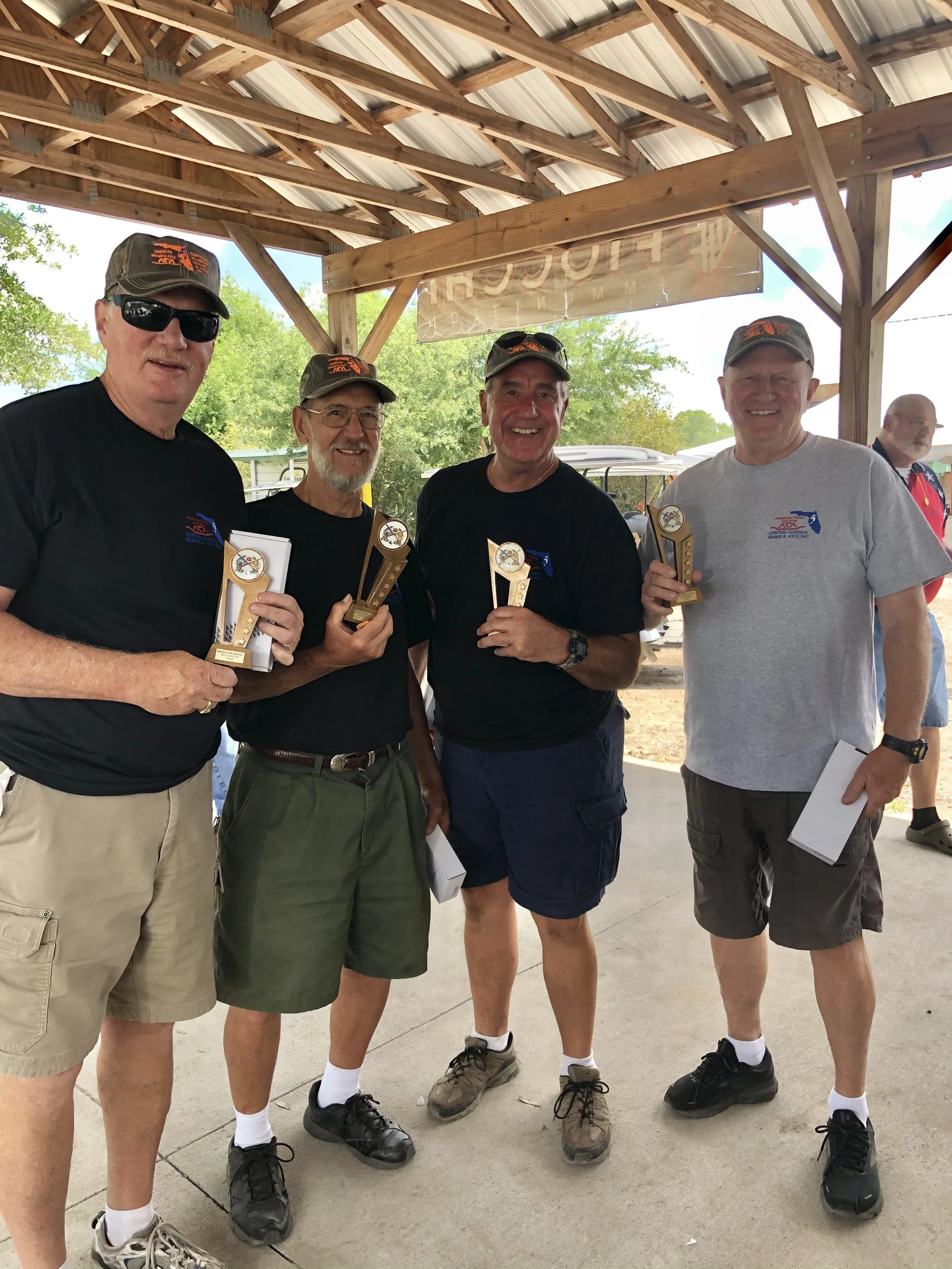 Four men standing under a wooden shelter, holding trophies and smiling at the camera, with trees and a golf cart in the background.