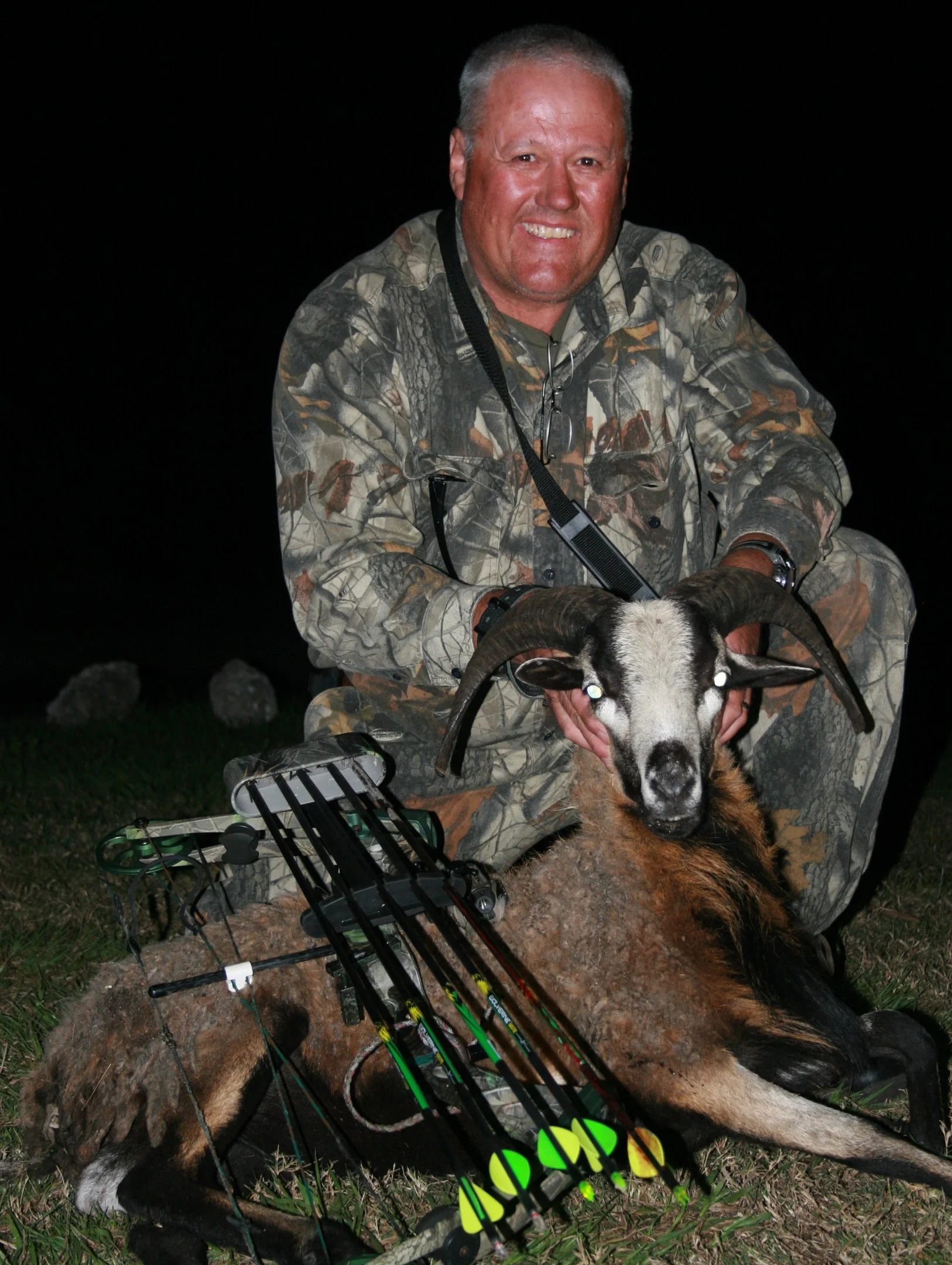 Man in camouflage hunting outfit kneeling beside a freshly hunted goat with hunting gear and a bow on the ground during nighttime.