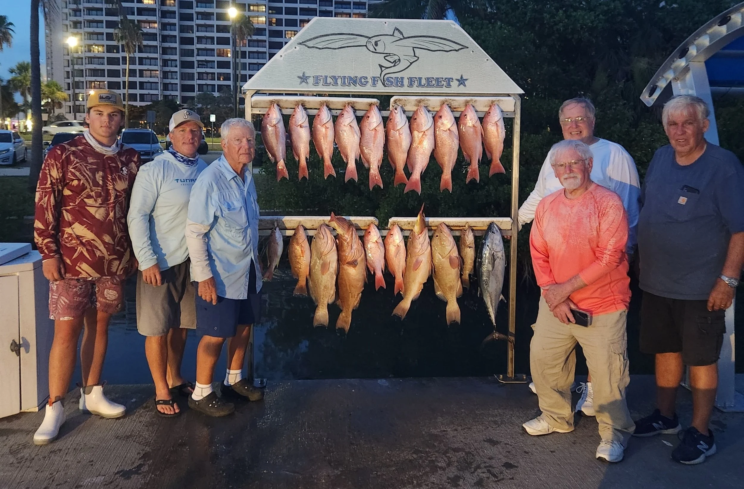 Group of seven men standing next to a display of fish caught during a fishing trip, with a sign that reads "Flying Fish Fleet" and a cartoon flying fish illustration, in an outdoor urban setting during dusk.