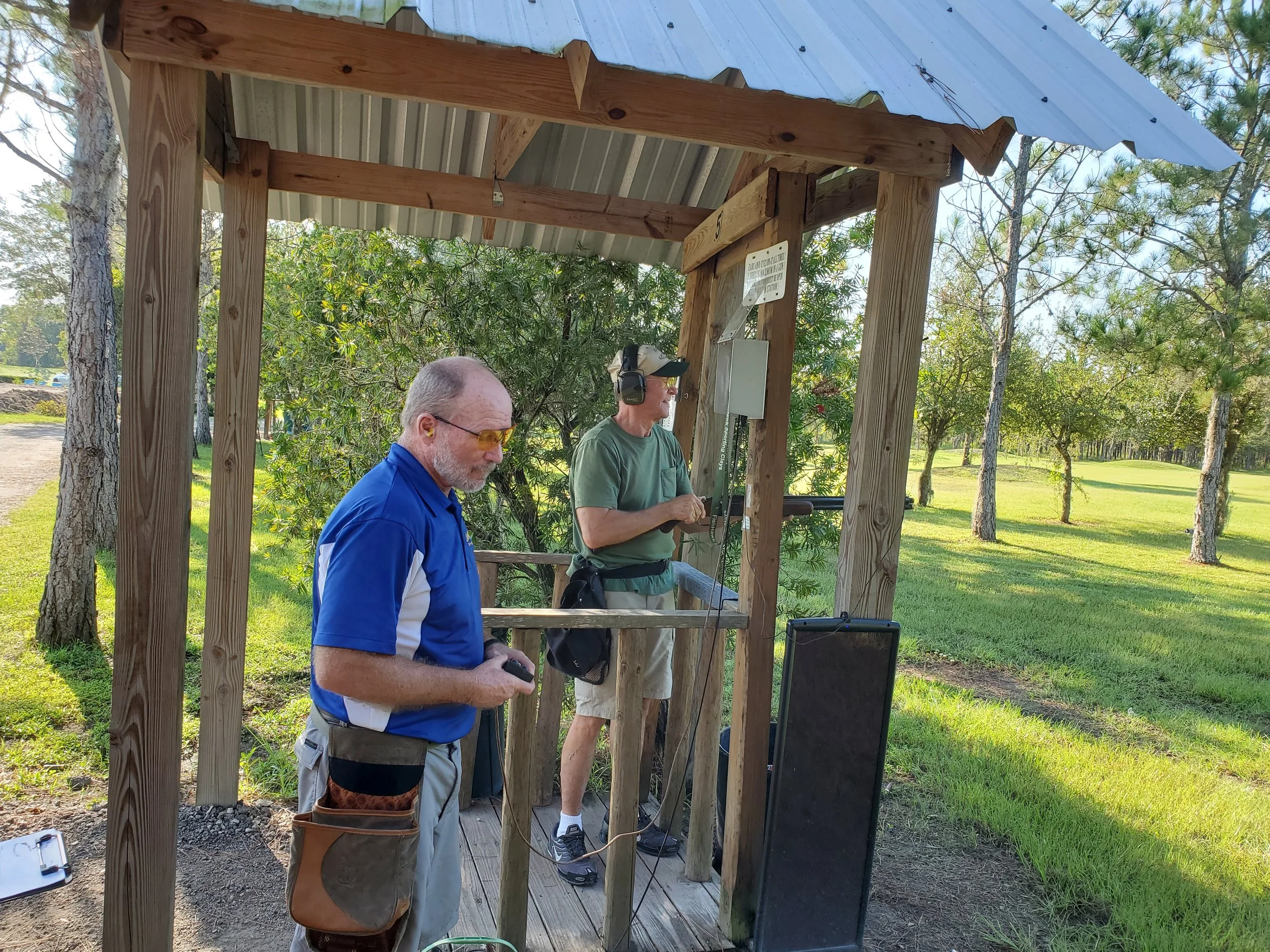 Two men at a shooting range under a wooden shelter. One man is wearing a blue shirt and glasses, holding a device in his hand. The other man, wearing a green shirt and earmuffs, appears to be aiming a firearm. The background shows green grass, trees,