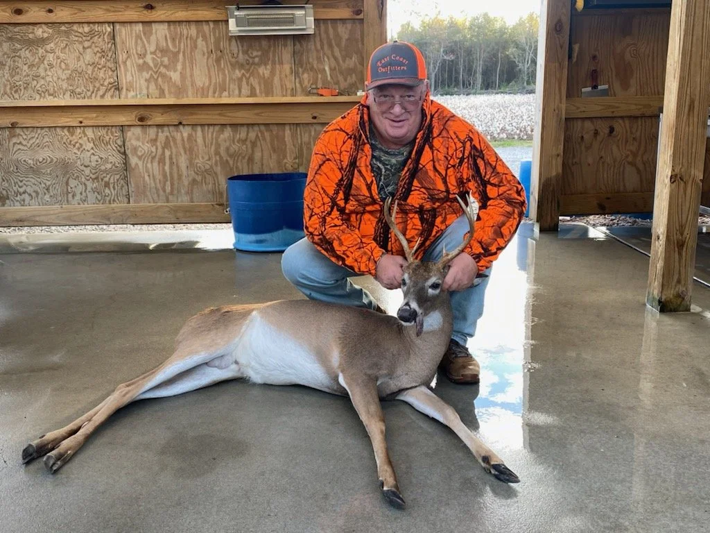 A man wearing an orange hunting jacket and a cap kneels next to a large dead deer laying on the ground inside a wooden shelter, smiling at the camera.