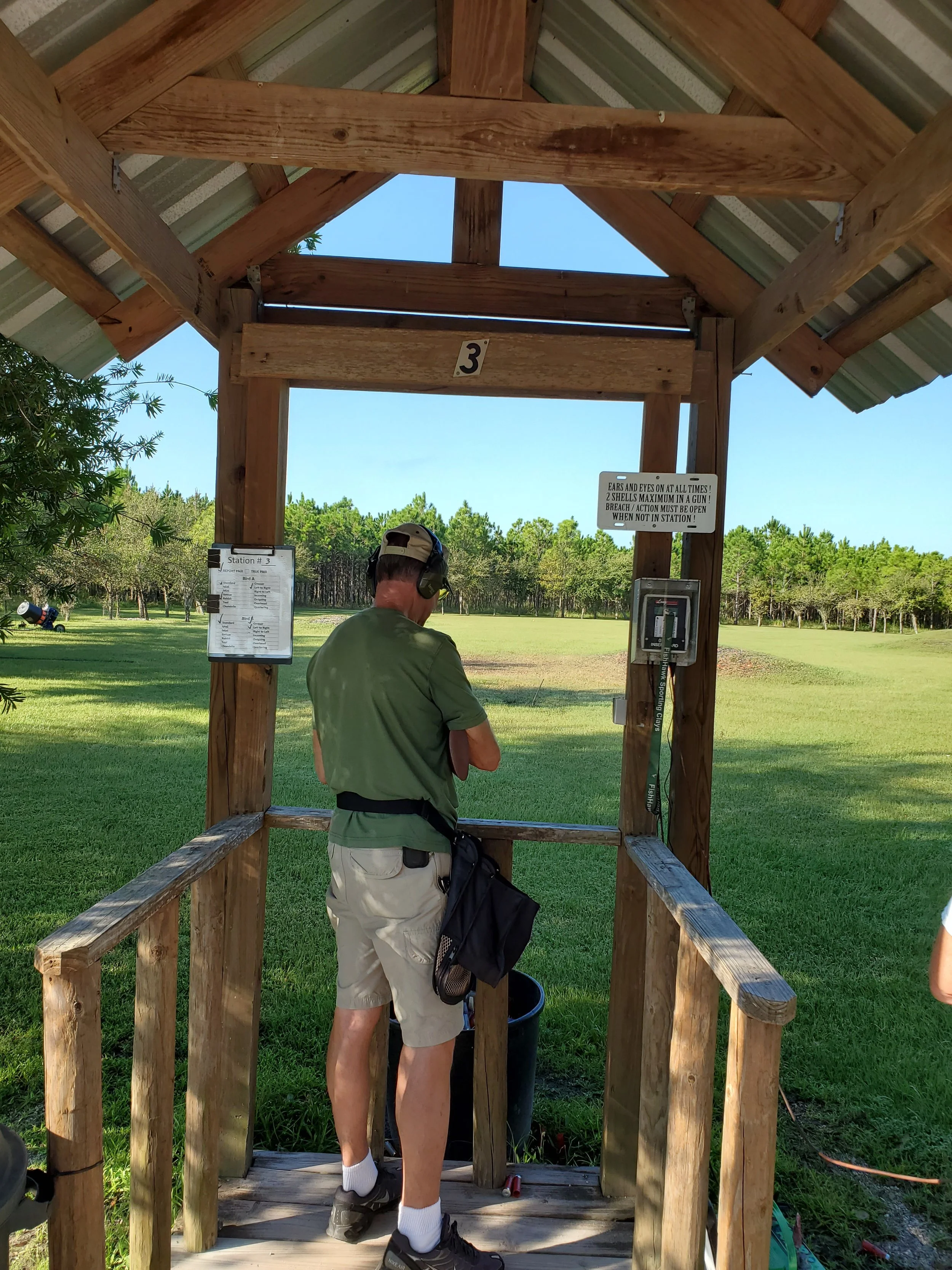 A person standing in a wooden shooting station at a shooting range, wearing headphones and ready to shoot.