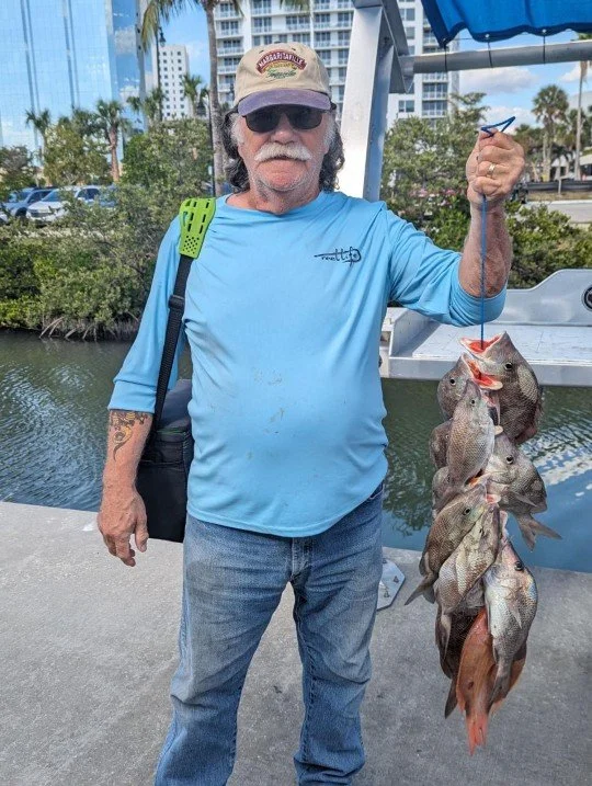An older man with a hat, sunglasses, and a mustache stands on a dock holding up a string of freshly caught fish. He is wearing a light blue long-sleeve shirt, jeans, and has a black shoulder bag. In the background, there are trees, a waterway, a boat