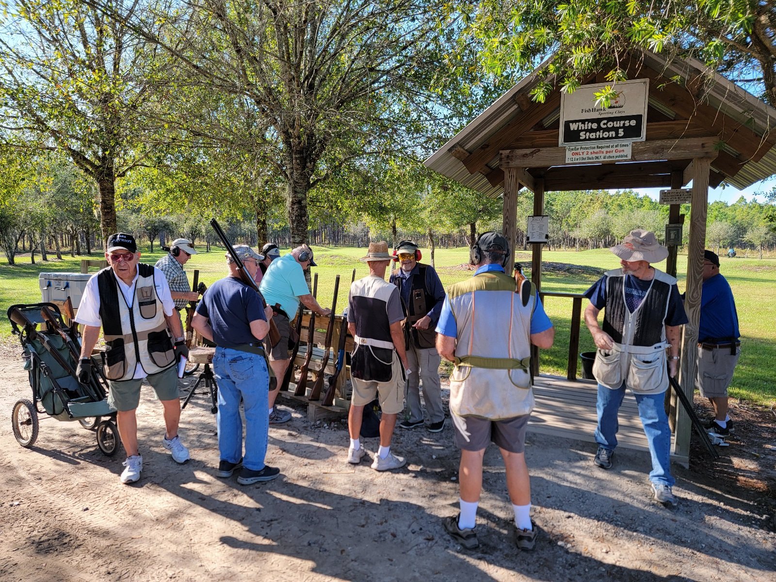 A group of elderly men at a shooting range practicing target shooting with shotguns, under a wooden shelter with trees and green grass in the background.