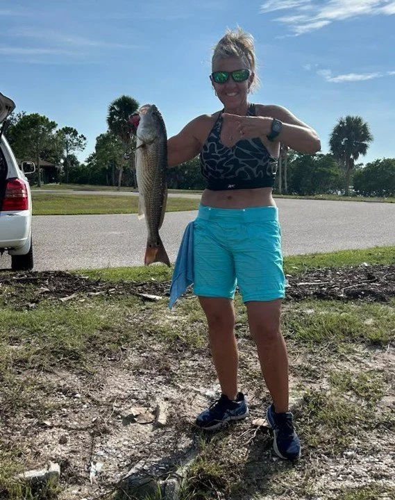 Woman standing outdoors, holding a freshly caught fish, wearing sunglasses, a patterned sports bra, and bright blue shorts, with a blue towel draped over her waist, near a parking lot with trees and a partly sunny sky in the background.
