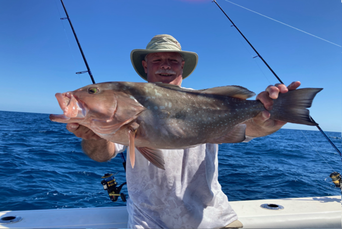 Man wearing a wide-brimmed hat holding a large fish on a boat, with a fishing rod and ocean in the background.