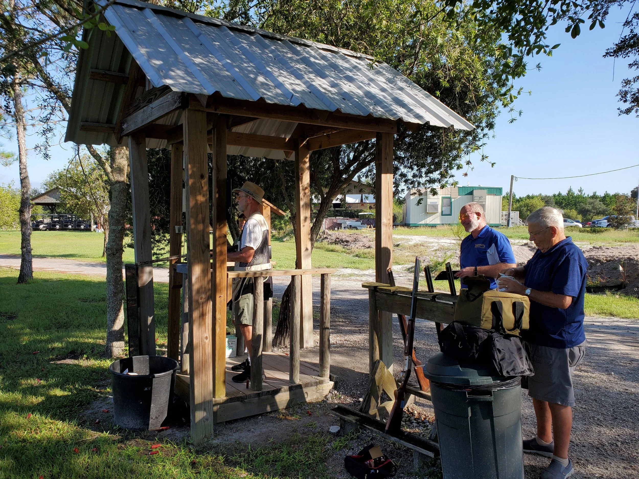 Three men at a small outdoor shooting range, two are standing and one is standing behind a wooden table, with one aiming a handgun at a target. There is a small wooden shelter, trees, and a trailer in the background.