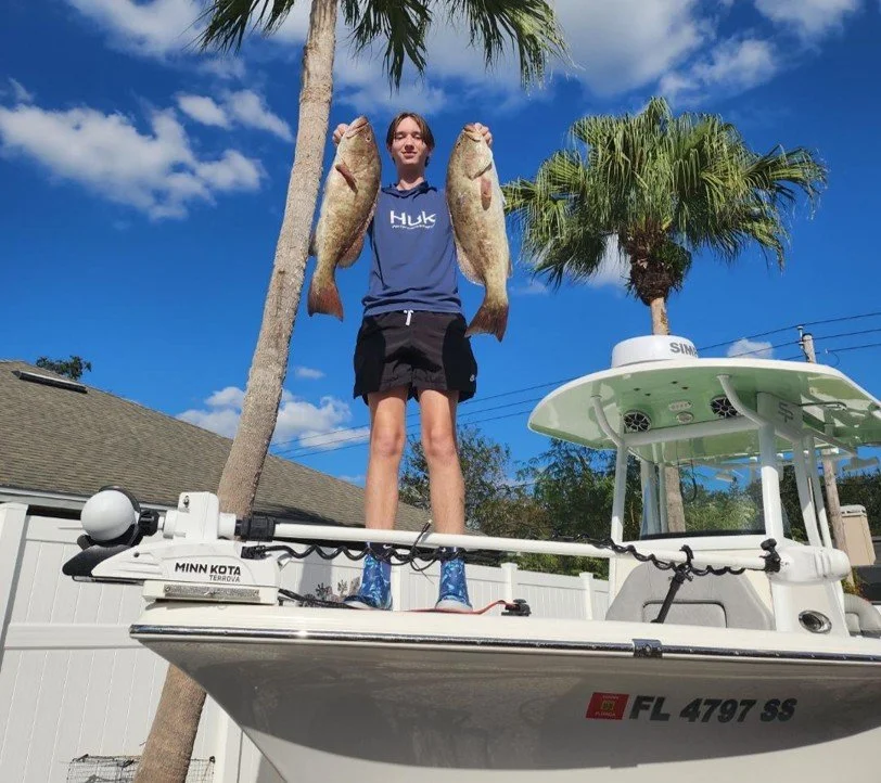 A person standing on a boat holding two large fish, with palm trees and a blue sky in the background.