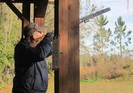 Person shooting a firearm at an outdoor range, wearing protective gear, under a wooden shelter with trees in the background.