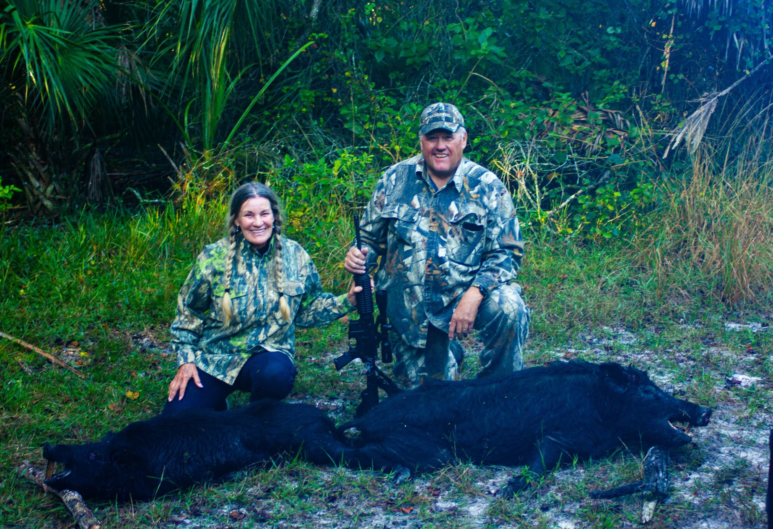 Two hunters, a woman and a man, in camouflage clothing kneeling beside two dead wild boars in a forested area, with the woman smiling and the man holding a rifle.