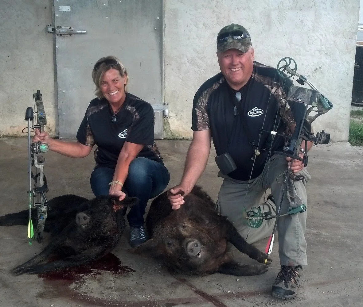 A man and woman kneeling on the ground, smiling and holding bows, next to two dead pigs, in front of a concrete wall.