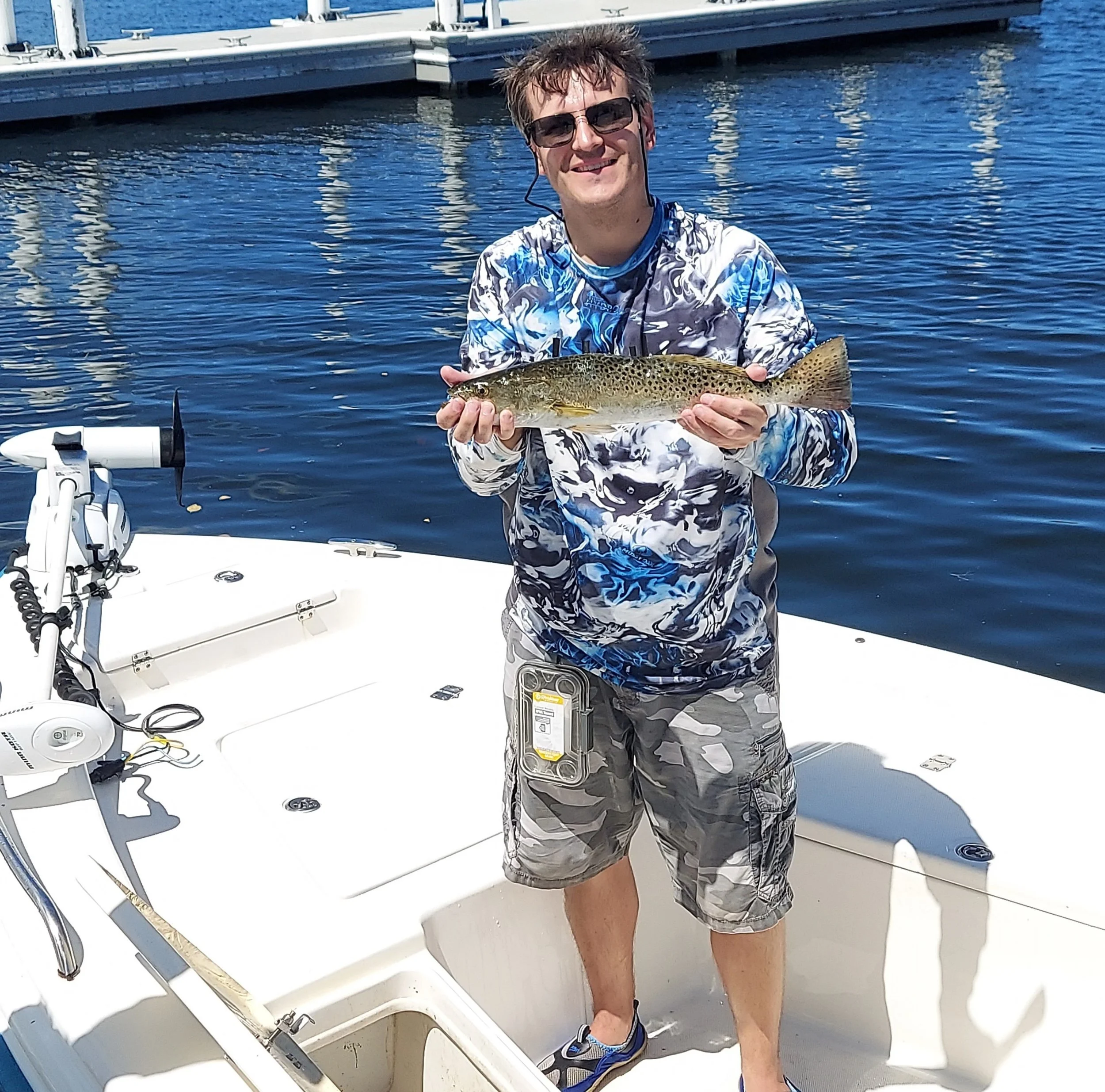 A man wearing sunglasses and a camouflage shorts, holding a fish on a boat. The boat is on water with other boats in the background.