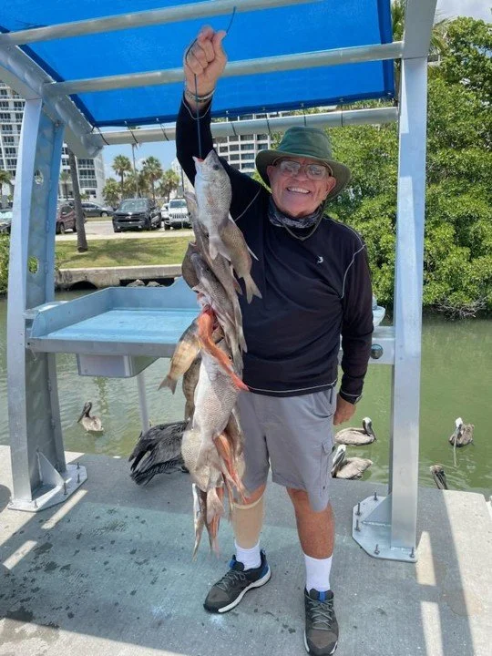 Smiling elderly man wearing a hat, sunglasses, and outdoor clothing, holding a string of fish over a dock near a waterway, with ducks swimming in the background.