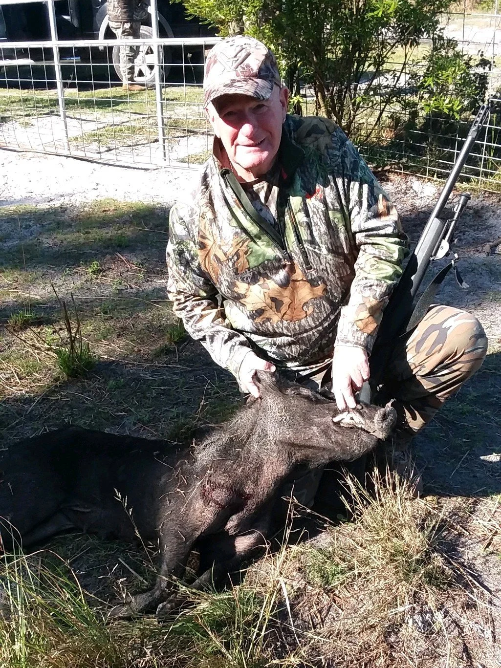 Man dressed in camouflage clothing crouching outdoors, holding a large, dead wild boar on the ground. There is a metallic fence and greenery in the background.