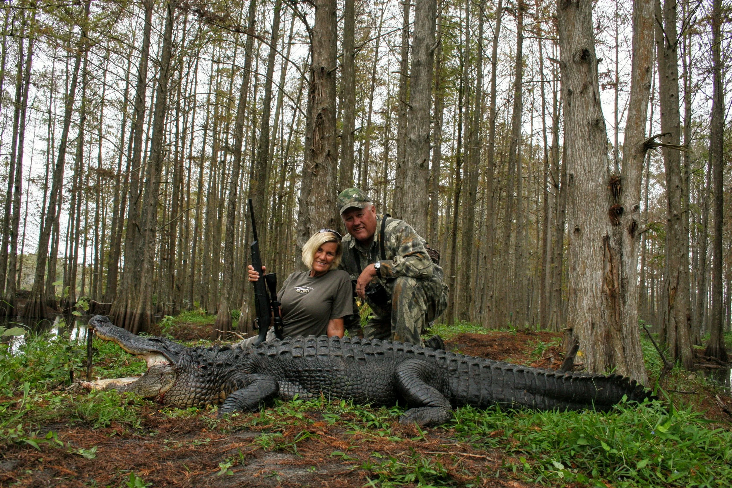 A woman and man in camouflage clothing posing with a large alligator on the ground in a wooded swamp area, with trees and water in the background.