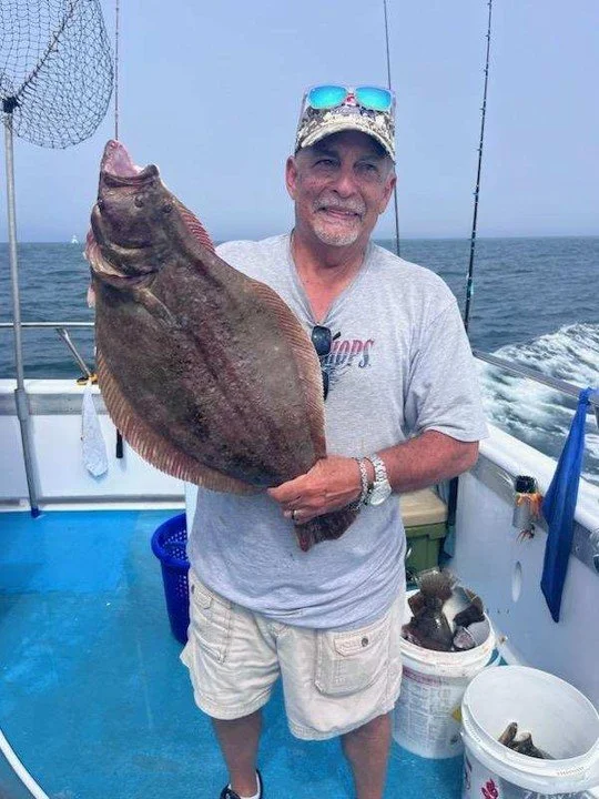 Man on a boat holding a large fish he caught, with fishing rods in the background and buckets of fish around him.