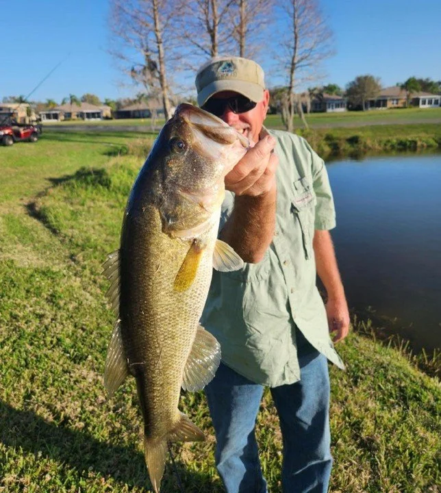 Man in hat and sunglasses holding a large bass fish near a pond on a sunny day.