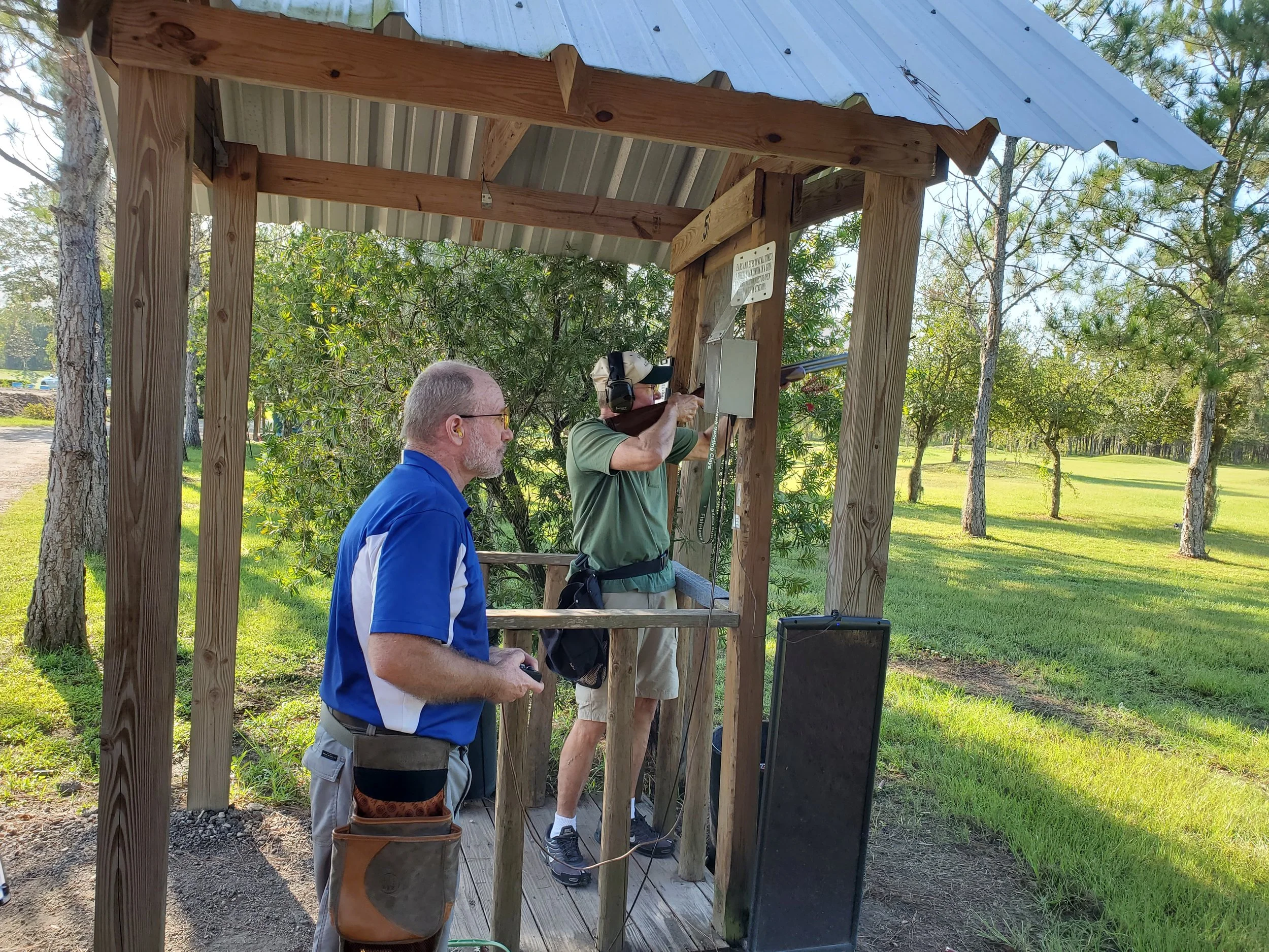 Two men at a shooting range, one aiming a firearm and the other observing, in a wooden shelter surrounded by trees.