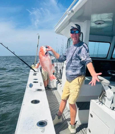 Man standing on a boat holding a large fish he caught, with fishing rods visible in the background and the ocean extending to the horizon.