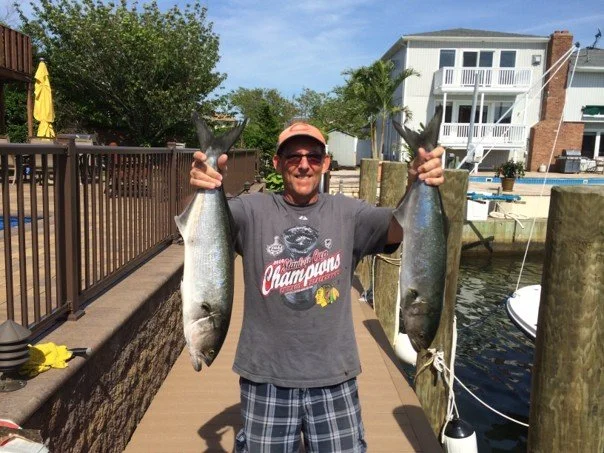 Man holding two large fish, standing on a dock by the water with houses and trees in the background.