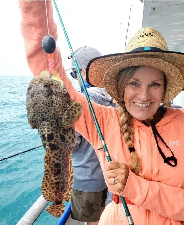 Smiling woman holding up a leopard spotted fish she caught while fishing on a boat. She is wearing a wide-brimmed straw hat and an orange hoodie.