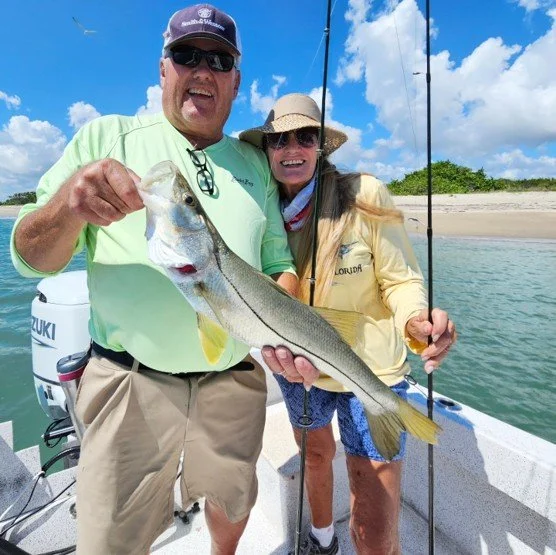A man and woman holding a large fish on a boat with a beach and blue sky in the background.