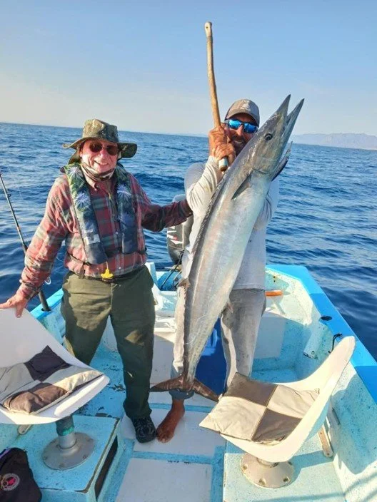 Two people on a boat holding a large fish. One person is smiling, wearing sunglasses, a hat, and outdoor gear, while the other person is holding the fish, wearing sunglasses, a cap, and casual clothing.
