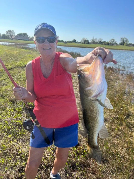 A woman wearing sunglasses, a blue cap, a pink sleeveless top, and blue shorts, smiling and holding a large fish she caught with a fishing rod near a water body on a sunny day.