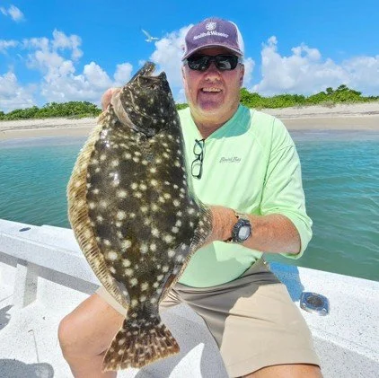 Man in a green shirt and sunglasses holding a large spotted fish on a boat at the beach with blue sky and clouds