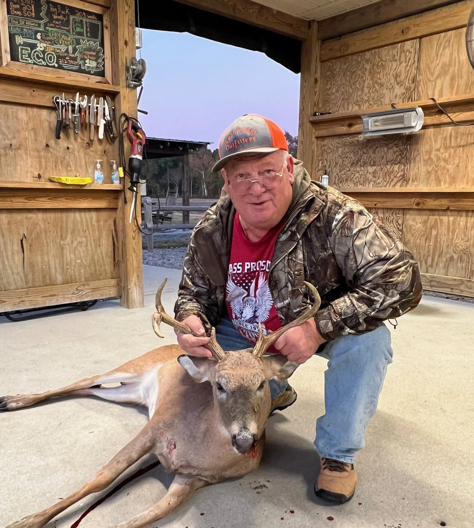 A man wearing camouflage jacket, red shirt, and cap kneeling inside a wooden structure, holding the antlers of a dead deer with a bloodstain on its neck, laying on the floor.