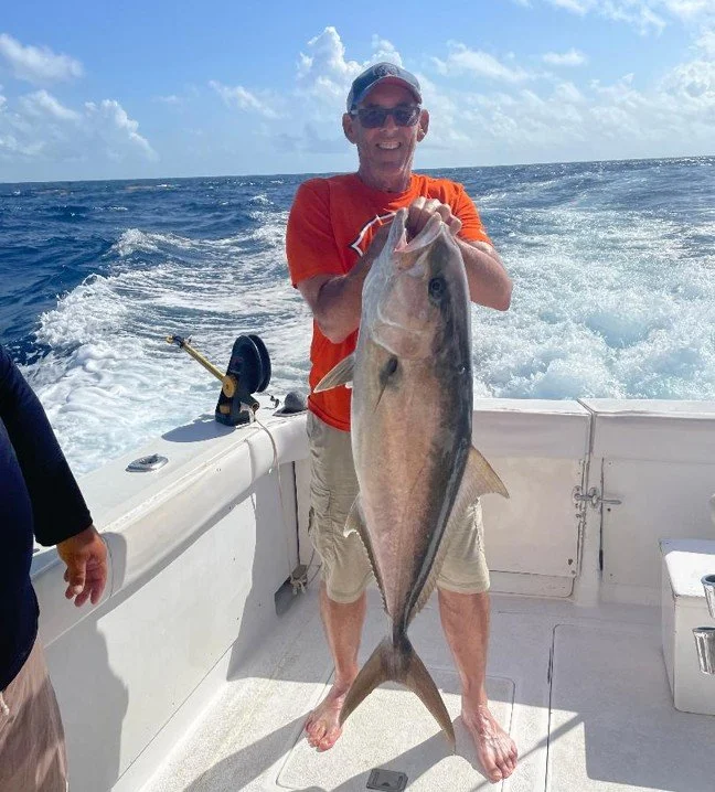 A man wearing a red shirt, beige shorts, sunglasses, and a cap holding a large fish on a boat in the ocean, with water and cloudy sky in the background.