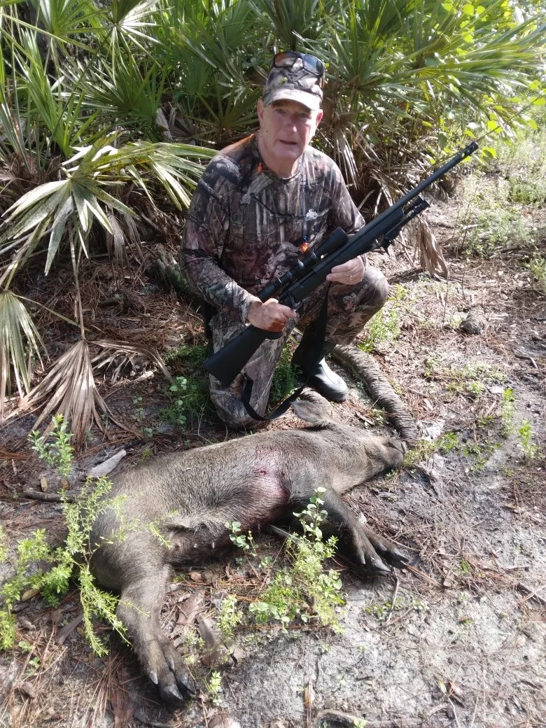 A man dressed in camouflage outdoors kneeling next to a dead wild boar, holding a rifle, with dense green foliage in the background.