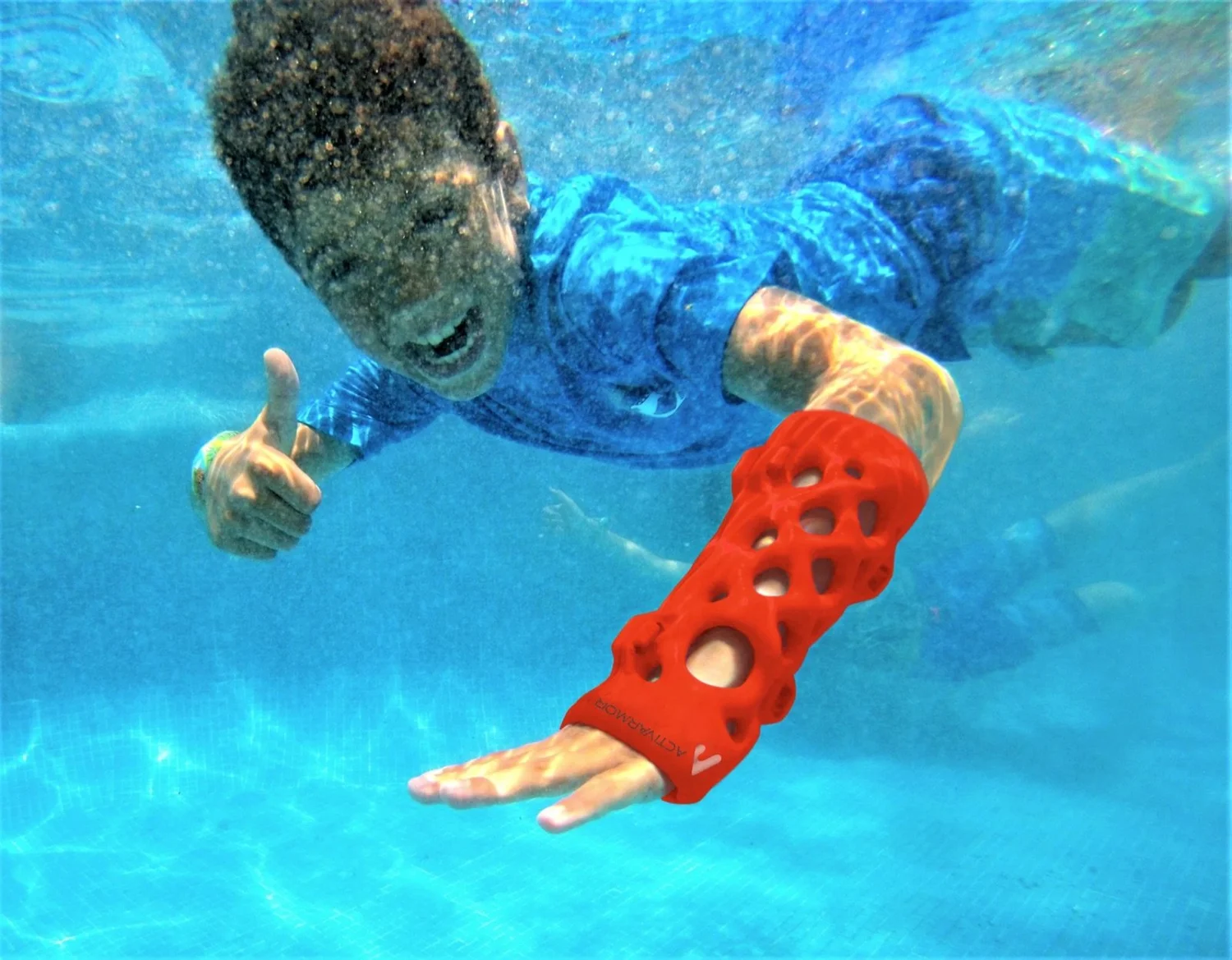 Niño nadando bajo el agua con su yeso ActivArmor rojo, haciendo señal de pulgar arriba