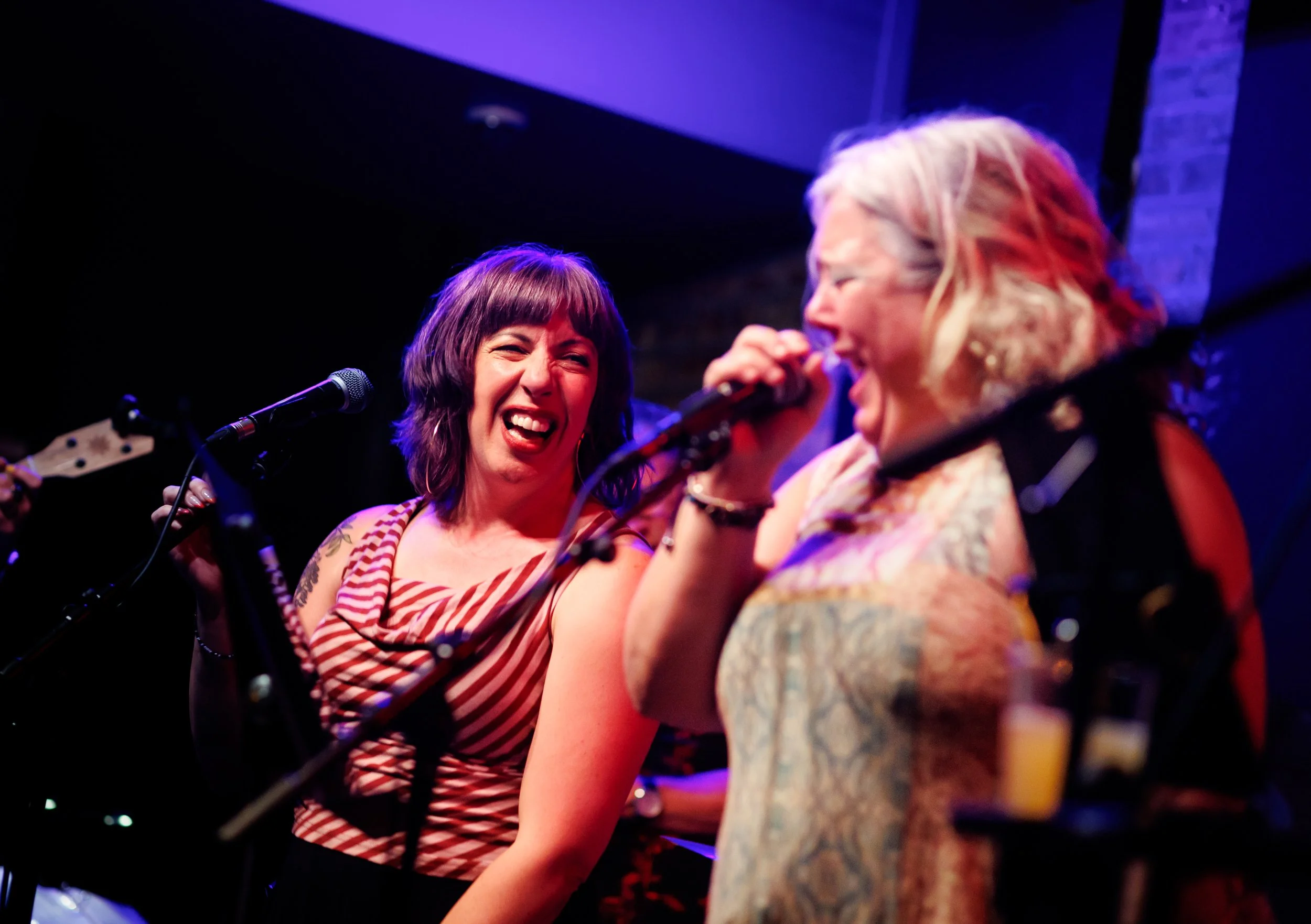 Two women singing and playing music on stage at a live event, smiling and laughing, with microphones in front of them.