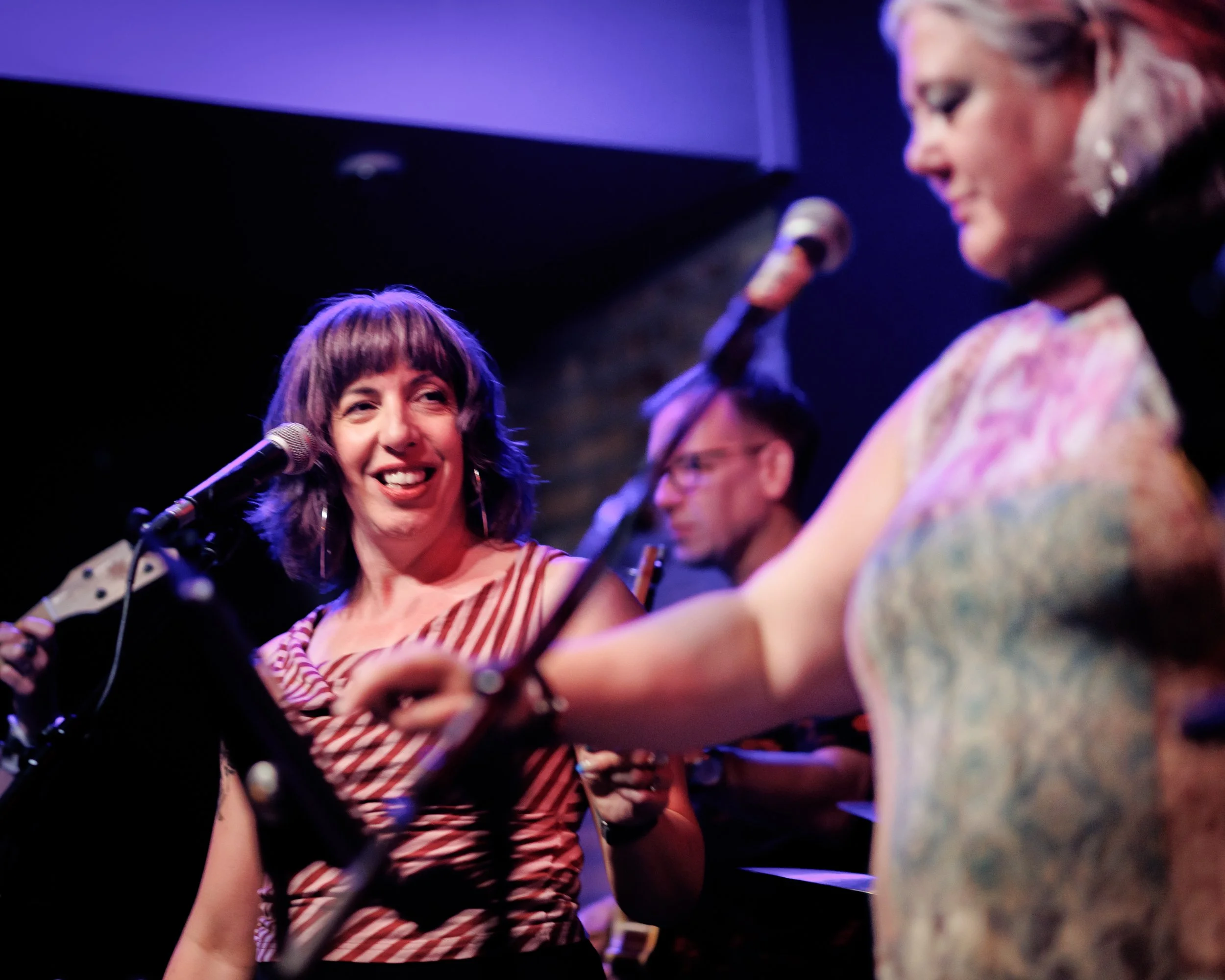 Three women on stage during a musical performance, with microphones and musical instruments, smiling and engaging with each other.
