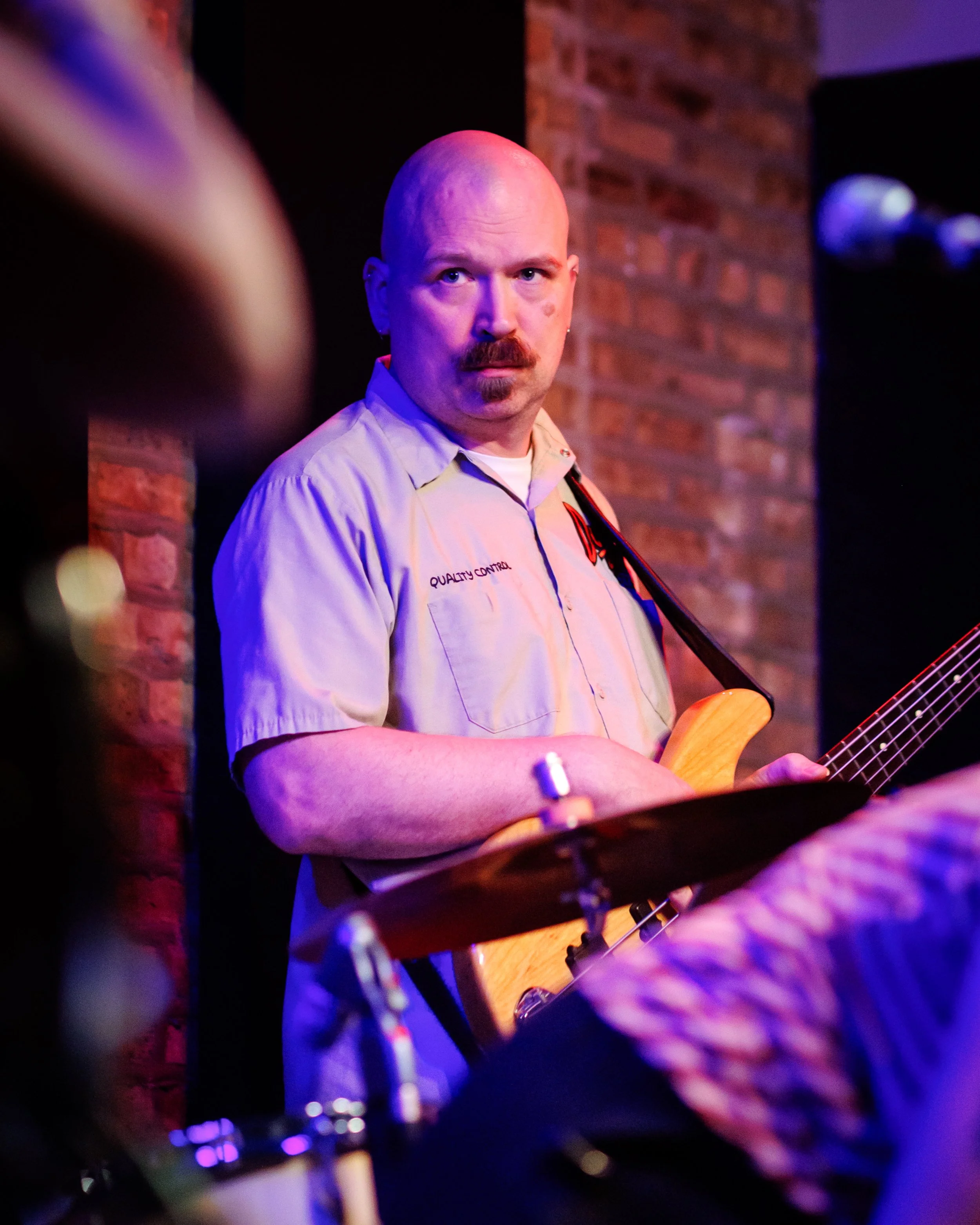 A man with a bald head, mustache, wearing a beige shirt, holding a guitar on stage in a nightclub or bar with brick walls and purple lighting.