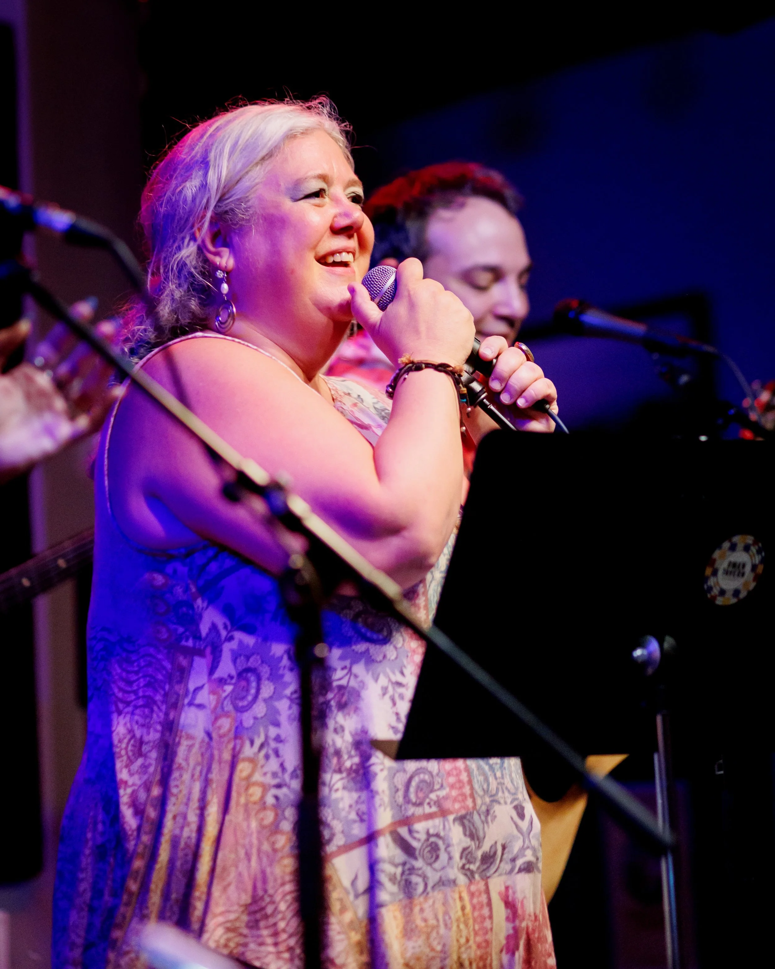 A woman singing into a microphone during a live music performance at an indoor venue, with a man playing guitar behind her.