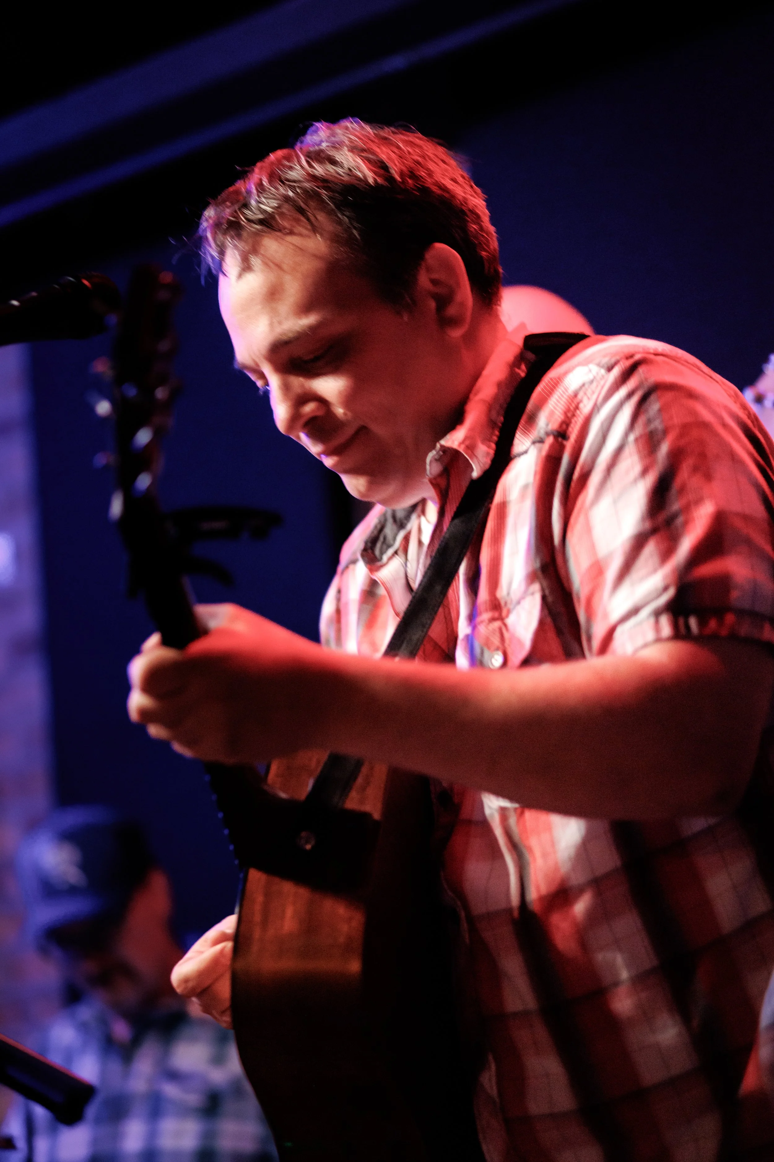 A young man playing an acoustic guitar, wearing a red and white plaid shirt, in a dimly lit setting with stage lights.