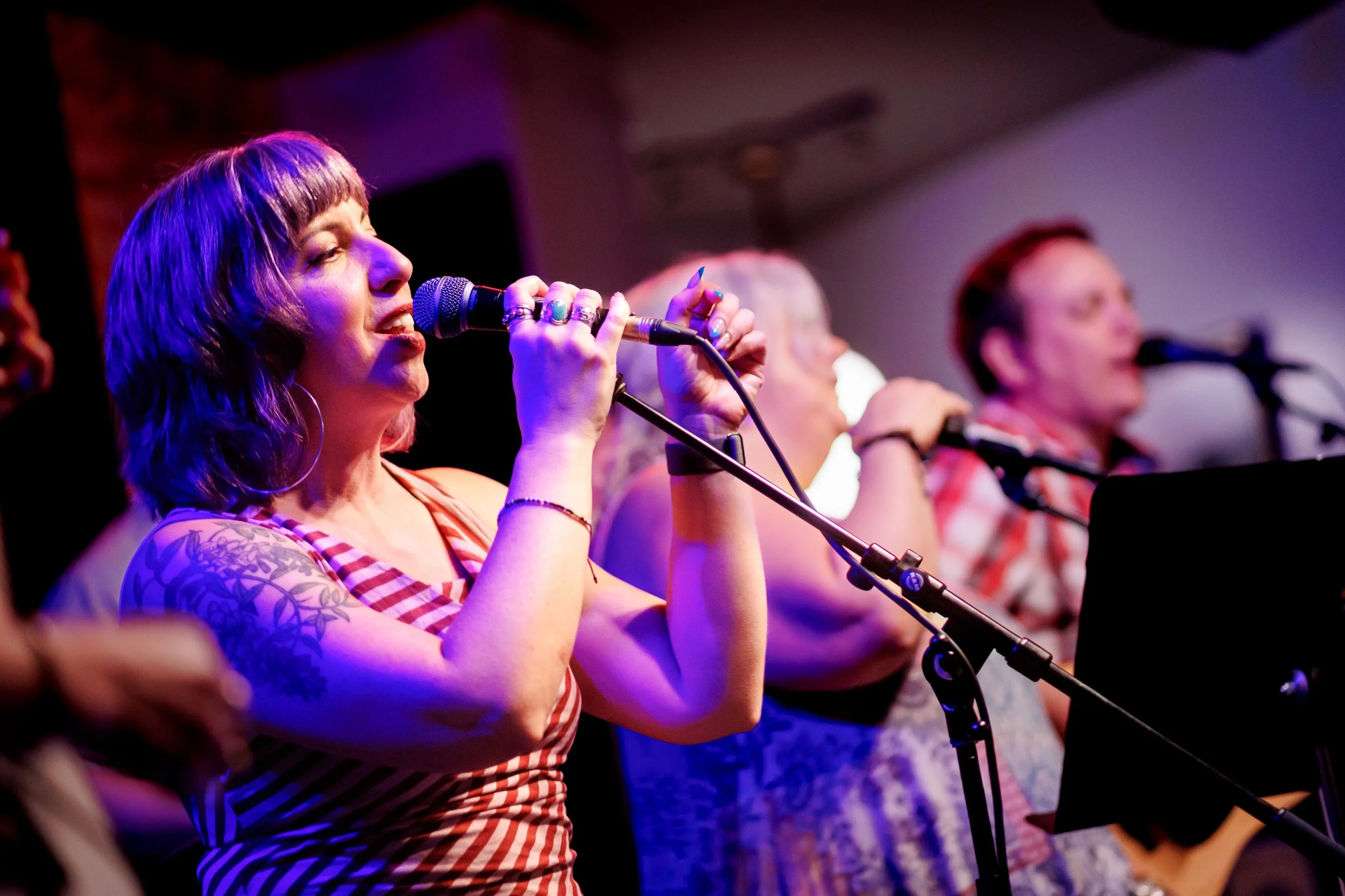 Three women singing into microphones on stage, with vibrant lighting and blurred background.